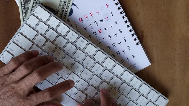 Close up top down view of Asian male finger typing on modern computer wireless keyboard on calendar and US dollar money around