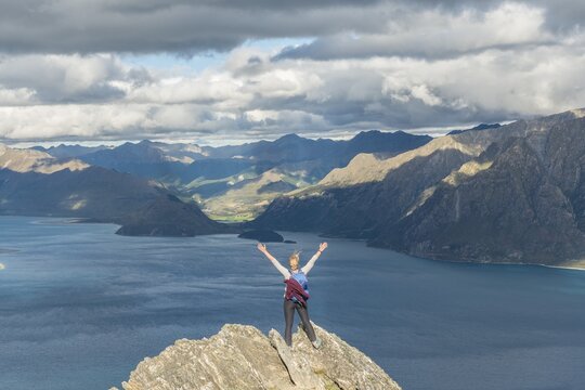 Female hiker on a rock, stretching arms in the air, Lake Hawea and mountain landscape, Isthmus Peak, Otago, South Island, New Zealand