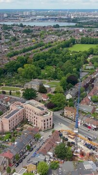 Drone flight toward Lloyd Park and William Morris House, Walthamstow, London. Forward aerial over forest road with Victorian terraced houses on the right and city skyline beyond. Vertical format.