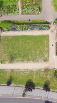 Drone view over Lloyd Park and William Morris House, Walthamstow, London. Top-down forward aerial reveals lawns, paths, circular plaza and nearby road traffic in sunshine. Vertical format.