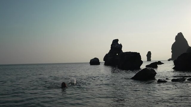 Rock formation, sea stacks, swimming woman enjoying the cold water near dramatic, jagged coastal cliffs under a wide, moody sky.