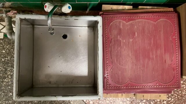 Rusty iron foot sink under faucet, set against green ceramic tiles, exposed pipes and valve, stained basin floor with clogged