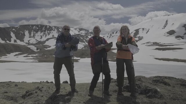 Group of happy senior friends having fun, taking off hats and dancing together on frozen lake in mountains. Elderly travelers. RAW flat profile uncorrected footage.
