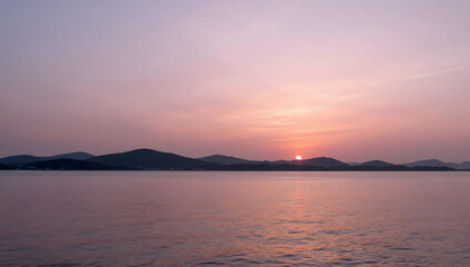 Fototapeta premium Serene Sunset Over Water: A breathtaking sunset paints the sky with hues of pink and orange, casting a soft glow over a calm body of water and a distant, silhouetted mountain range.