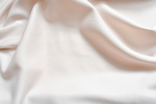 Cream white silk fabric texture with soft waves and folds top view. Detailed close up of gentle vanilla satin cloth background with smooth drapery and stitched edge