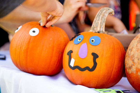 Two pumpkins decorated with stick on foam faces and googly eyes at a no carve Halloween craft activity as a hand adds a sticker, capturing kid friendly autumn fun and a knife free decorating idea.