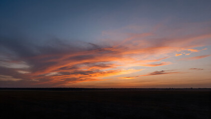Fototapeta premium Dramatic Sunset Over Open Field: The sky explodes with vibrant hues of orange and pink as a fiery sunset paints the clouds above a vast, dark expanse of a field.
