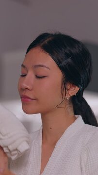 Young Asian woman in white robe holding soft towel near face after cleansing skincare routine, calm hygiene portrait in bathroom wellness beauty relaxation indoor light Vertical