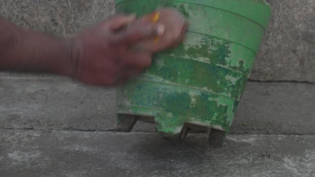 A person is cleaning a green cement flowerpot by hand using a coconut coir brush. In which a green plant is planted. One hand holds the edge of the pot while the other scrubs the damaged surface.