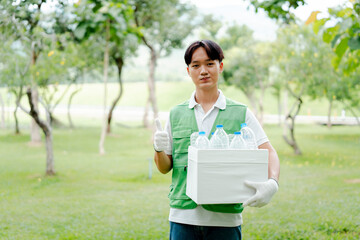 Fototapeta premium Young asian man volunteer wearing green vest and gloves. Holding a box of plastic bottles for recycling. Proudly giving thumbs up gesture in a park