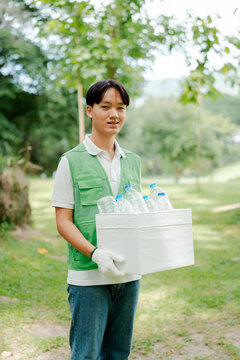 Young asian man volunteer wearing a green vest and white gloves, collecting plastic bottles in a white container at a park, contributing to environmental protection and recycling efforts