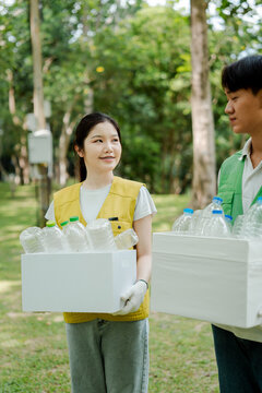 Young asian female and male volunteers collecting and carrying boxes full of plastic bottles in a public park, focusing on environmental recycling and sustainability efforts