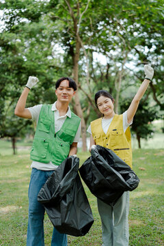 Young asian male and female volunteers wearing vests and gloves. Holding full trash bags. Posing and celebrating a successful park cleanup