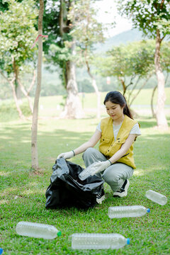Young asian volunteer collecting plastic bottles into a trash bag, cleaning park environment, promoting recycling and sustainable waste management in nature