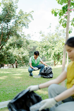 Young asian volunteers collaborate in a park cleanup, picking up trash and plastic into black bags on the grass, promoting environmental protection and community teamwork