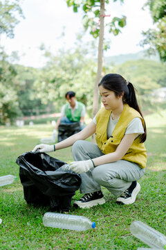 Young asian person volunteering and collecting plastic bottles in a black garbage bag at a park cleanup event, contributing to environmental protection and sustainability efforts