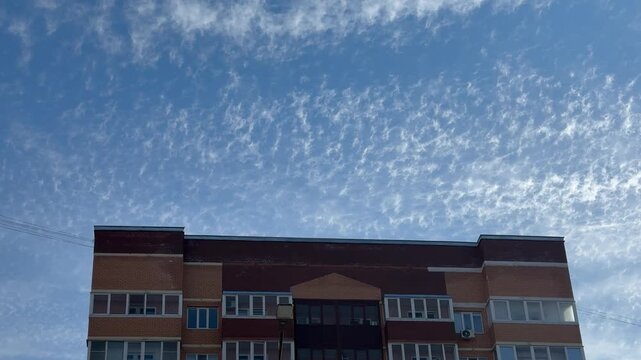 Brick apartment tower under blue sky, sunlit red facade with repeating balcony rows and window patterns, wispy clouds, crisp urban