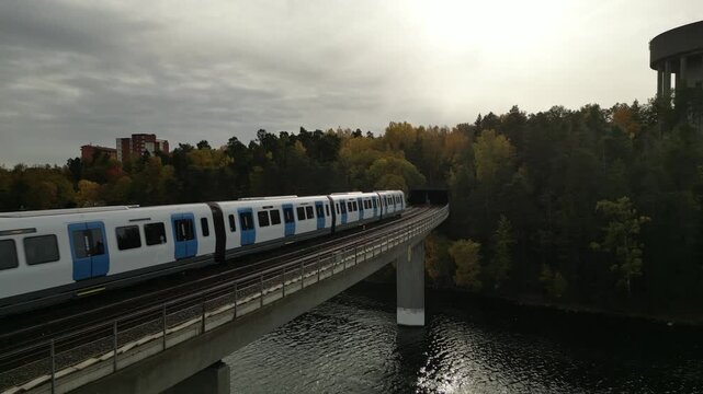 Cinematic aerial drone footage of a modern passenger train traveling across a bridge above water and entering a colorful autumn forest. The drone gradually gains altitude, revealing vibrant fall folia