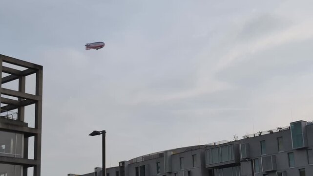 Weather balloon  airship moves slowly over city streets, hovering above tall buildings. People below look up at the strange sight in the sky.