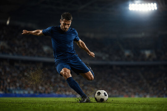 Professional Soccer Player in Blue Uniform Kicking Ball on Grass Pitch in Stadium at Night. Dynamic Action Shot of Male Athlete Striking Football during Match.