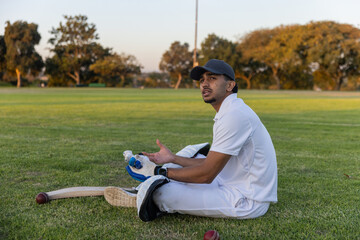 Fototapeta premium Indian male cricketer sitting on field wearing pads with bat and red ball nearby, copy space