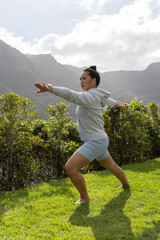 Fototapeta premium Standing lunge pose balancing on well kept grassy lawn, with trimmed hedge and mountain ridge