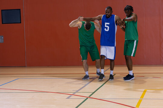 Diverse male basketball players helping limping teammate across gym court with jerseys and shoes