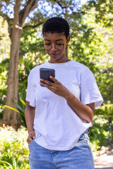 Fototapeta premium African woman standing in sunny park wearing white tee and jeans wearing glasses holding smartphone