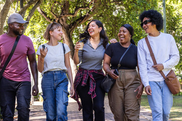Fototapeta premium Diverse friends walking on shaded park path holding coffee cup and phones, wearing shades and bags