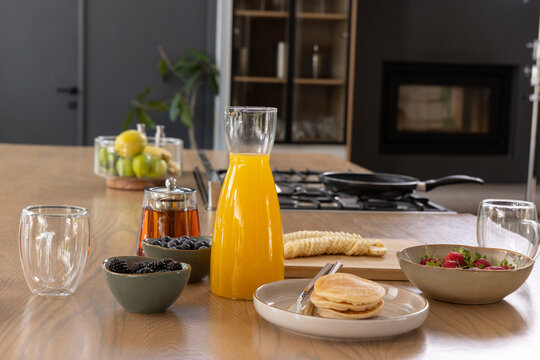 Clear glass carafe of orange juice sitting centered on wooden island, with pancakes and berries