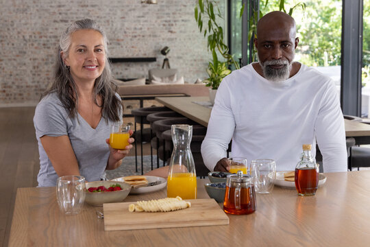 Diverse senior couple sitting at home table holding glasses of orange juice, with bowl of berries