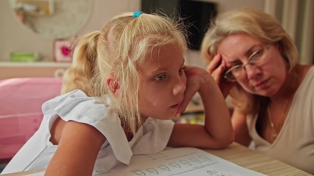 Schoolgirl leaning on hand while using laptop for homework, mother assisting nearby. Tired during remote learning at home