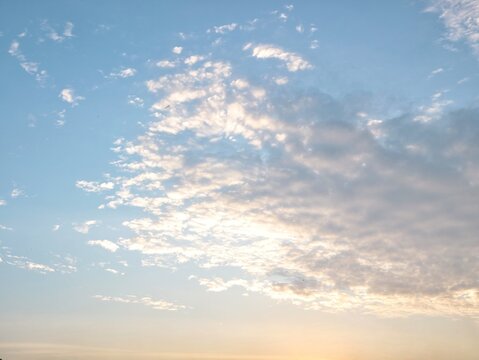 A scenic view of textured white altocumulus clouds scattered across a bright blue sky, illuminated by the warm, golden glow of the sun during a late afternoon sunset or early sunrise