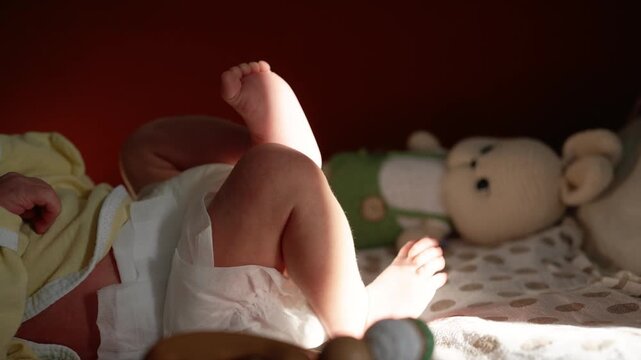 Adorable newborn boy in a diaper playfully kicking his tiny feet and legs, illuminated by a warm sunbeam, while resting on a changing table next to his favorite teddy bear