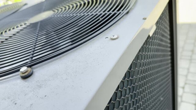 An air conditioning unit operates outside a residential home. The system cools the house, producing the loud sound of a spinning fan and the roar of a motor.