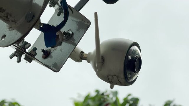 A white security camera monitors an outdoor area during a rainstorm. Raindrops fall continuously onto the wet plastic and metal frame. This creates a gentle tapping sound.