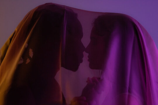 Multiracial couple kissing under transparent veil at studio. Privacy and discretion concept. 