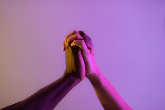 Hands of anonymous multiracial couple holding each other firmly against lilac studio background