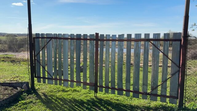 Weathered metal gate with vertical stainless strips, chain link fences flanking sides, spring grass carpeting ground, clear blue