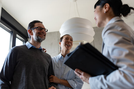 Happy diverse couple laughing with a real estate agent during an apartment tour. Smiling multicultural clients looking at paperwork, discussing a property lease, home purchase, or mortgage deal