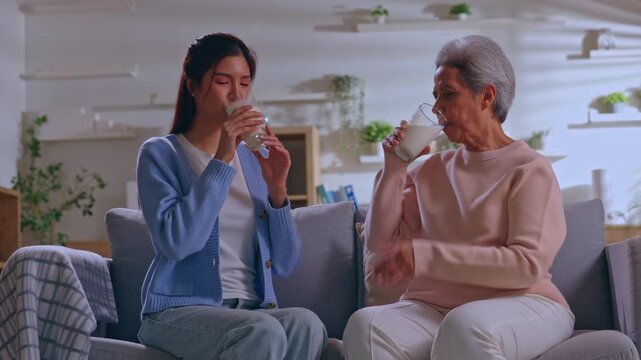Asian woman caregiver with senior woman holding milk glass on sofa at home, family conversation, warm drink, smiling care in cozy living room with plant shelf and comfort