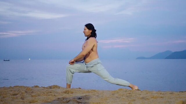 Man Doing Yoga Lunge on sandy Seashore at Dusk. Shirtless man in light pants holds a deep yoga lunge on a rocky shoreline with calm water, a pastel evening sky, and distant landforms in the background