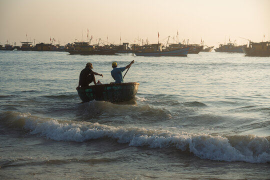Fishermen guide a traditional basket boat through shallow waves near Tam Tien coast, Da Nang, Vietnam at sunrise.