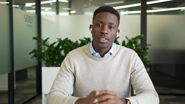A young african american man in a beige sweater, blue collared shirt and earpiece, uses natural gestures at a white minimalist desk in a bright co-working space, bathed in natural light. Concept of