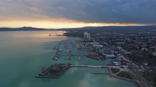 In Balatonf&uuml;red, moored sailing boats line the Lake Balaton marina beside the waterfront promenade and nearby buildings, viewed from a drone in soft evening light under a cloudy sky.