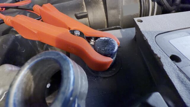 A hand attaches a red clamp to a car battery under the hood. This simple action brings a comforting sense of relief and hope during an unexpected vehicle breakdown.
