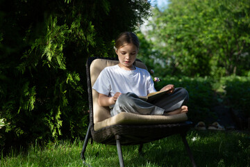 Obraz premium Happy adorable smiling child girl reads a book sitting on a chair in the summer garden near the house. Happy ghild having fun outdoors. Generation Alpha. copy space. 