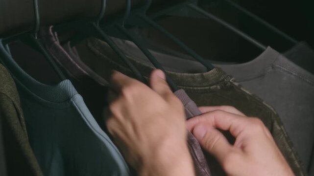 Close up man hands browsing through selection colorful t shirts on hangers, trying decide what to wear and choosing perfect outfit from organized home closet. Man choosing clothes from wardrobe closet