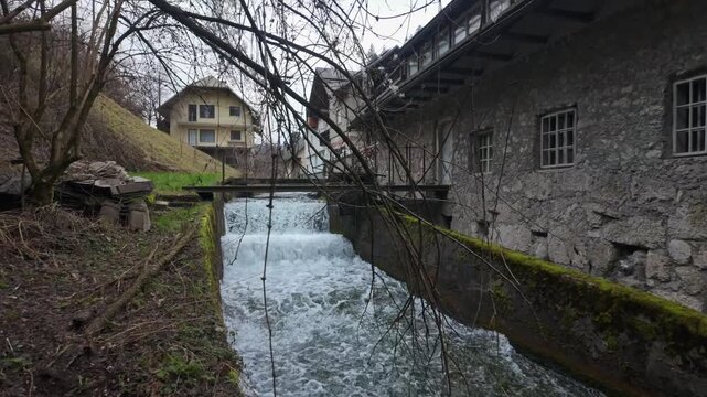 In Bled, Slovenia, a small winter waterfall cascades through a forested village setting with bare trees and traditional houses beneath an overcast sky, captured in slow motion.