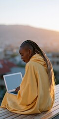 Obraz premium Woman in a yellow hoodie seated on a wooden deck using a laptop at sunset with mountain and city views for remote work background and cozy outdoor scene